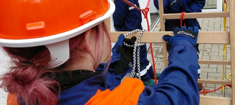 Kinder in Feuerwehrkleidung üben Knoten mit Seilen an einer Leiter